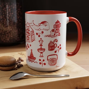 May include: A white ceramic mug with a red interior and handle. The mug features red line drawings of a house, whale, lighthouse, and other coastal elements. A macaron, coffee beans, and a spoon are on a wooden surface.