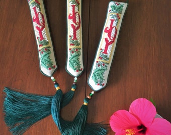 May include: Three white fabric ornaments with green and red embroidery spelling "JOY" with green tassels hanging from each. A pink hibiscus flower is in the bottom right corner.