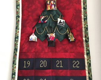 May include: A red and green Christmas advent calendar with a quilted tree design. The tree has 12 pockets for ornaments and a calendar with the numbers 19 through 24. The calendar is signed "Sew Exquisite".