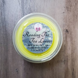 May include: A round, clear plastic container holding a bright yellow scented soy wax shot. The label reads "Reading The Tea Leaves" with additional text indicating it is vegan and hand-poured in Florida, with a net weight of 1.5 oz / 42.5 g.