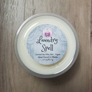 May include: A round, clear container holding a creamy white wax melt. The label reads "Laundry Spell" with additional text indicating it's a scented soy wax shot, vegan, hand-poured in Florida, and weighs 1.5 oz.
