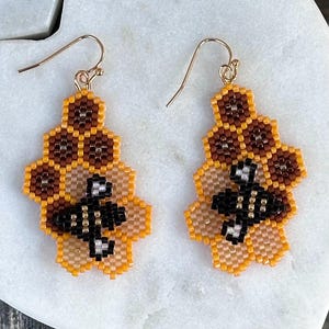 May include: A pair of handmade beaded earrings. The earrings feature a honeycomb design with a bee in the center. The colors include shades of brown, yellow, black, and white. The earrings have gold-colored ear wires.