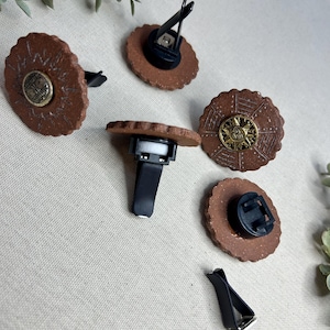 May include: Several brown, circular car air fresheners with decorative designs and black clips. Some have gold-colored centerpieces. The air fresheners are arranged on a neutral-colored surface, with greenery in the background.