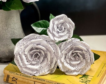 May include: Three handcrafted paper roses made from sheet music with black musical notes on white paper, featuring green leaves. The roses rest on a yellow book titled Mammoth Series No. 10 Robbins Mammoth Collection of Modern Piano Music.