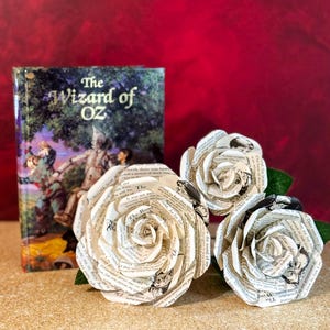 Op de afbeelding: Drie witte papieren rozen gemaakt van pagina's uit het boek "De Tovenaar van Oz". De rozen zijn op een bruine ondergrond geplaatst met het boek op de achtergrond.