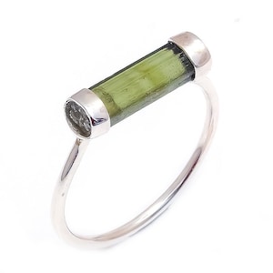 Puede incluir: Un anillo de plata con una piedra preciosa de turmalina verde engastada en un engaste de bisel. La piedra preciosa tiene una forma rectangular larga y está engastada horizontalmente en la banda.