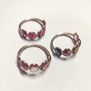 May include: Three silver rings, each featuring a band of colorful gemstones. Two rings display pink stones, while the third ring has a combination of pink, blue, and yellow stones. The rings are set against a white background.