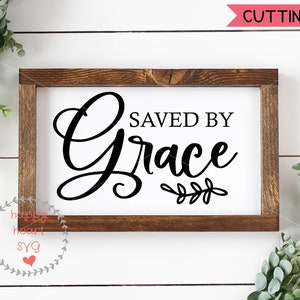 Puede incluir: Un letrero blanco con un marco de madera marr&oacute;n, que presenta el texto "Saved by Grace" en letras cursivas negras con una peque&ntilde;a rama de hojas debajo.