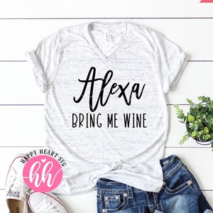 May include: A white t-shirt with a black graphic that says "Alexa Bring Me Wine". The shirt is laying on a white wooden surface with a pair of white sneakers and blue jeans.