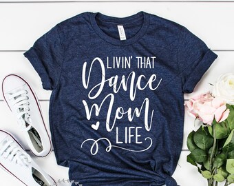 May include: A navy blue t-shirt with the text "Livin' That Dance Mom Life" in white cursive lettering. The shirt is on a white wooden surface with white sneakers and pink roses.