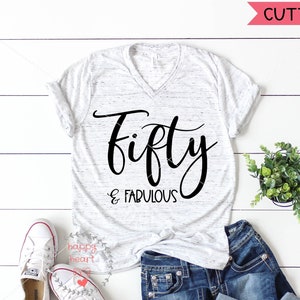 May include: A white t-shirt with a black graphic that says "fifty & fabulous". The shirt is on a white wooden surface with a pair of white sneakers, blue jeans, and a small potted plant.