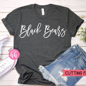 May include: A dark gray t-shirt with the text "Black Bears" in white script lettering. The shirt is laid flat on a white wooden surface with a pair of white sneakers and a pair of denim shorts.