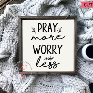 May include: A white wooden sign with black text that reads "Pray More Worry Less". The sign is sitting on a white knitted blanket with a cup of coffee next to it.