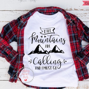 以下が含まれることがあります： 黒い文字で「The Mountains Are Calling And I Must Go」と書かれた白いTシャツ。テキストは2つの黒い山のシルエットで囲まれています。