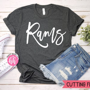 May include: A dark gray t-shirt with the word "Rams" written in white cursive script. The shirt is laid flat on a white wooden surface with a pair of white sneakers and a pair of blue denim shorts.