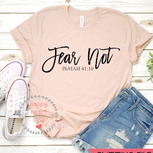 Puede incluir: Una camiseta rosa con el texto "Fear Not" en letras cursivas negras y el texto "ISAIAH 41:10" en negro debajo. La camiseta est&aacute; sobre una superficie de madera blanca con un par de zapatillas blancas y unos pantalones cortos de mezclilla azul.