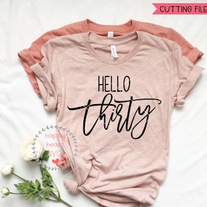 May include: A pink t-shirt with the text "HELLO thirty" in black script font. The shirt is folded and laid flat on a white surface.