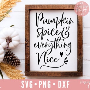 Puede incluir: Un letrero de madera con fondo blanco y texto negro que dice "Pumpkin Spice & everything Nice" con un pequeño corazón y un diseño de espiral. El letrero está enmarcado en un marco de madera marrón oscuro.