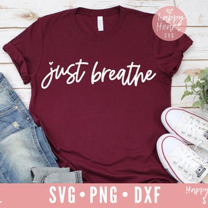 Op de afbeelding: Een maroon t-shirt met de tekst "just breathe" in witte letters. Het shirt ligt plat op een wit houten oppervlak met een paar witte canvas schoenen en een blauwe jeans.