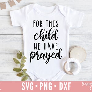 Op de afbeelding: Witte baby romper met zwarte tekst die "Voor dit kind hebben we gebeden" luidt.