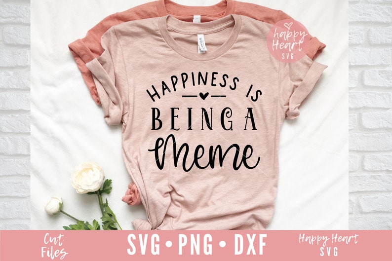May include: A pink t-shirt with the text "Happiness is being a meme" in black lettering. The shirt is folded and laid on a white surface with a white flower.