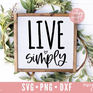 Könnte beinhalten: Ein Holzschild mit dem Text "LIVE simply" in schwarzen Buchstaben. Das Schild ist von grünem Laub umgeben.