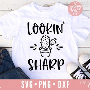 Puede incluir: Camiseta blanca con texto negro que dice "Lookin' Sharp" con un gráfico de cactus. La camiseta está sobre una superficie de madera blanca con un par de pantalones vaqueros azules y zapatillas negras.