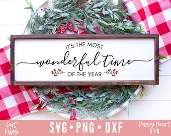 May include: A wooden sign with a white background and brown frame. The sign reads "It's the most wonderful time of the year". The sign is decorated with small red berries and green leaves.