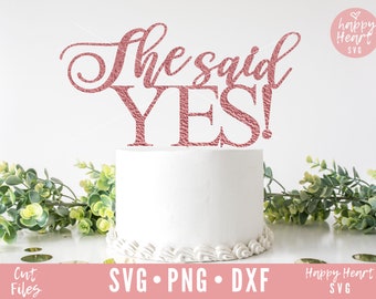 Puede incluir: Un pastel blanco con un adorno de purpurina dorada rosa que dice "She said YES!" El pastel está decorado con glaseado blanco y un borde blanco. El adorno está sobre un fondo blanco.