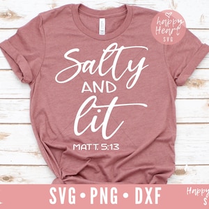 以下が含まれることがあります： 白い筆記体で「Salty and Lit」と書かれたピンクのTシャツ。メインテキストの下に「MATT. 5:13」というテキストが印刷されています。