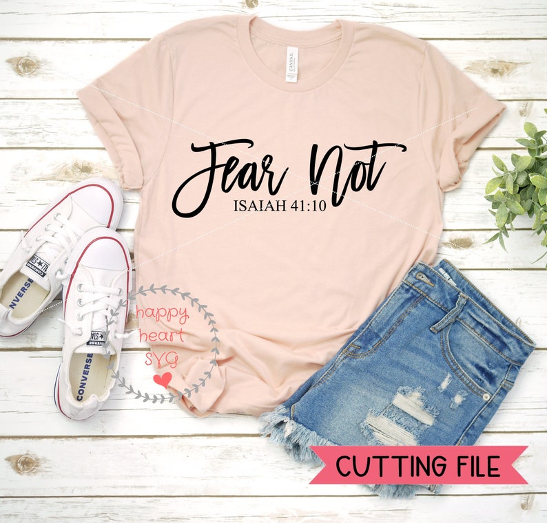 Puede incluir: Una camiseta rosa con el texto "Fear Not" en letras cursivas negras y el texto "ISAIAH 41:10" en negro debajo. La camiseta est&aacute; sobre una superficie de madera blanca con un par de zapatillas blancas y unos pantalones cortos de mezclilla azul.