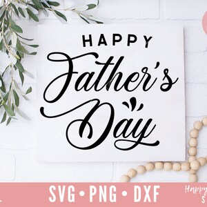 K&ouml;nnte beinhalten: Wei&szlig;er Hintergrund mit einem schwarzen Schild, auf dem in kursiver Schrift "Happy Father's Day" steht. Das Schild ist von Gr&uuml;npflanzen und Holzperlen umgeben.