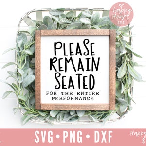 May include: A white sign with a brown frame, featuring the text "PLEASE REMAIN SEATED FOR THE ENTIRE PERFORMANCE". The sign is sitting in a white wicker basket filled with green leafy plants.