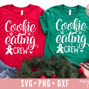 May include: Two red and green t-shirts with the text "Cookie eating crew" and a gingerbread man graphic. The shirts are laid out on a table with a green wreath and a plaid blanket.