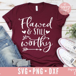 Op de afbeelding: Een maroon t-shirt met de tekst "Flawed & STILL worthy" in witte letters met twee kleine hartjes en een pijlgrafiek.