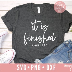 May include: A dark gray t-shirt with the text "it is finished" in white cursive lettering and the scripture reference "JOHN 19:30" below. The shirt is laid out on a white wooden surface with a pair of white sneakers and a pair of blue jeans.