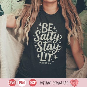 以下が含まれることがあります： 白いグラフィックが「Be Salty Stay Lit」と書いてある濃いグレーのTシャツ。テキストの周りに星があり、グラフィックの下に「Matthew 5:13-14」という文字があります。