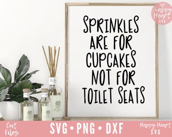 May include: A white sign with black text that reads "Sprinkles are for cupcakes not for toilet seats". The sign is framed in a light wood frame and is sitting on a table with a plant and bottles.