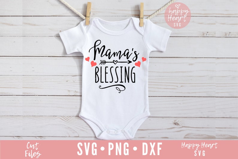 Op de afbeelding: Een witte baby romper met zwarte tekst die "Mama's Blessing" zegt met een pijl en een hart. Er zijn kleine rode hartjes rond de tekst.