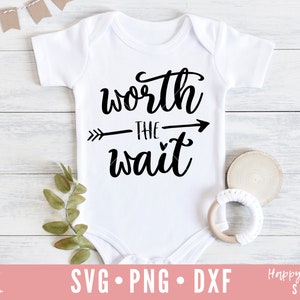 Op de afbeelding: Witte baby romper met zwarte tekst die "worth the wait" zegt met een pijl die naar rechts wijst. De romper ligt op een witte houten achtergrond.