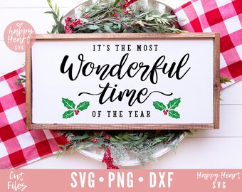 May include: A white wooden sign with the text "It's the most wonderful time of the year" in black cursive lettering. The sign is decorated with two green holly leaves with red berries on each side of the text.