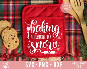 May include: Red oven mitt with the text "baking through the snow" in white. The oven mitt is on a red and black plaid surface with cookies and a wooden spoon.
