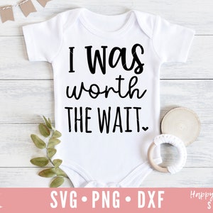 Op de afbeelding: Witte baby romper met zwarte tekst die "Ik was het wachten waard" zegt. De romper ligt op een witte houten achtergrond met een klein groen plantje en een houten ring met wit garen.