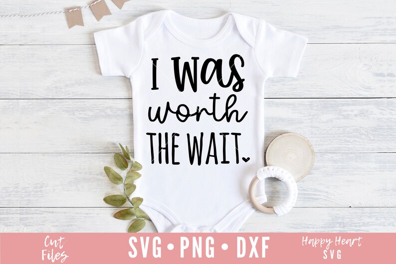 Op de afbeelding: Witte baby romper met zwarte tekst die "Ik was het wachten waard" zegt. De romper ligt op een witte houten achtergrond met een klein groen plantje en een houten ring met wit garen.