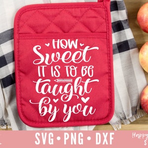 以下が含まれることがあります： 白地に「HOW Sweet IT IS TO BE taught by you」と書いてある赤いキルティングの鍋つかみ。鍋つかみは、黒と白のチェック柄の布が敷かれた木製の台の上に置かれている。