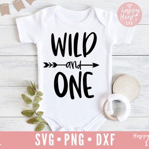 K&ouml;nnte beinhalten: Wei&szlig;er Strampler mit schwarzem Text, der "Wild and One" lautet, mit einem Pfeil, der nach rechts zeigt. Der Strampler liegt auf einer wei&szlig;en Holzoberfl&auml;che mit gr&uuml;nen Bl&auml;ttern und einem Holzring.