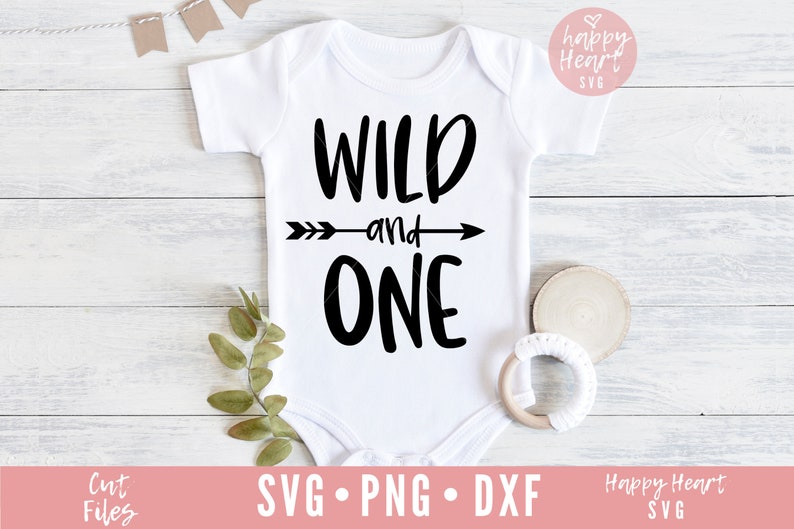 K&ouml;nnte beinhalten: Wei&szlig;er Strampler mit schwarzem Text, der "Wild and One" lautet, mit einem Pfeil, der nach rechts zeigt. Der Strampler liegt auf einer wei&szlig;en Holzoberfl&auml;che mit gr&uuml;nen Bl&auml;ttern und einem Holzring.