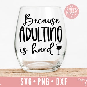 May include: A clear wine glass with the text "Because adulting is hard" and a small wine glass icon. The glass is sitting on a wooden surface.