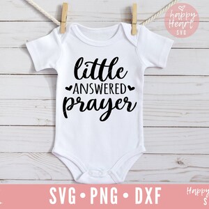 Op de afbeelding: Een witte baby romper met zwarte tekst die "little answered prayer" luidt, met een hartje aan weerszijden van het woord "answered".