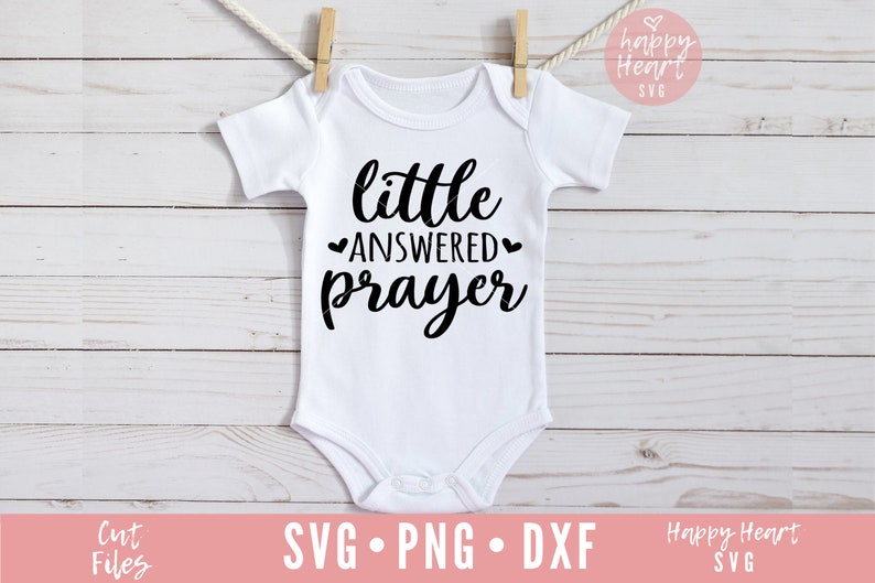 Op de afbeelding: Een witte baby romper met zwarte tekst die "little answered prayer" luidt, met een hartje aan weerszijden van het woord "answered".